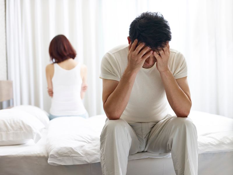 A man and a woman sit on opposite sides of a white bed with their heads in their hands.