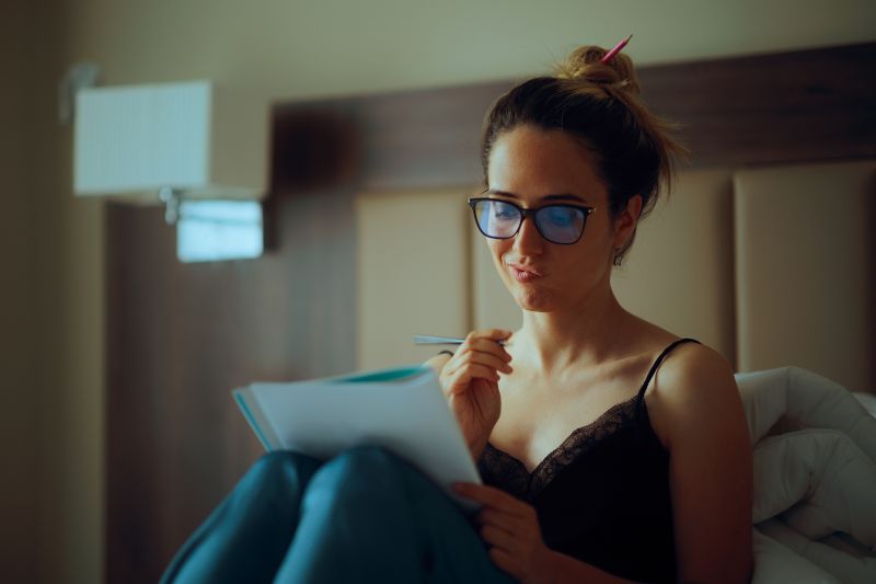 Woman in comfy clothes, glasses, and bun sitting on bed pondering over a journal